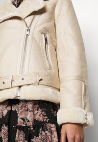 Beige shearling jacket with a wide collar, silver zippers, side pockets, and a belted waist; paired with a floral-patterned skirt.