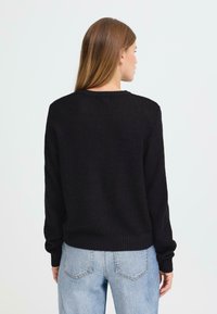 Svart strikket genser med teksturert mønster, rund hals, og lange ermer, kombinert med lyseblå jeans.