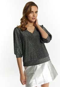 Glänzender schwarzer V-Ausschnitt-Pullover mit gepufften 3/4-Ärmeln, kombiniert mit einem metallisch silbernen Minirock, der eine glatte, reflektierende Textur aufweist.
