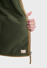Grøn fleecejakke indvendigt med lynlås, sandfarvet trim, sidelomme og T-KIT-logo. Blød tekstur med en ensfarvet og letvægtsdesign.