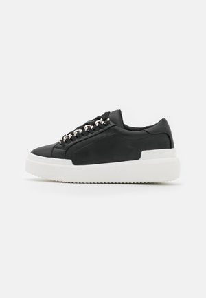 Czarny niski sneaker z grubą białą podeszwą i srebrnymi oczkami na sznurowadłach, pokazany od zewnętrznej strony na jednolitym białym tle.