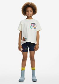 Camiseta de manga corta crema con parches impresos coloridos, combinada con pantalones cortos azules con diseños vibrantes. Botas de lluvia azules y calcetas amarillas.
