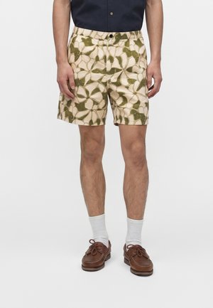Homme portant un short beige avec un motif abstrait de feuille verte, des chaussettes blanches hautes, des chaussures bateau en cuir marron et une chemise bleu marine rentrée.