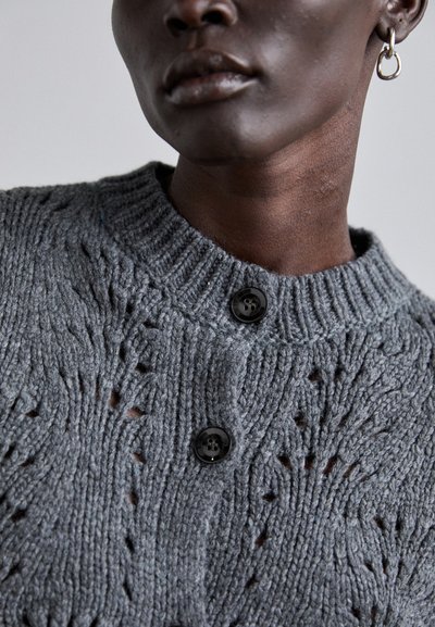 Grau gestrickte Strickjacke mit strukturiertem Muster, geripptem Kragen und zwei schwarzen Knöpfen auf der Vorderseite. Der Stoff wirkt weich und warm.