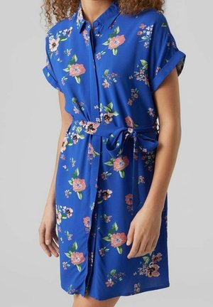 Blå kortærmet skjortekjole med pink og grøn blomsterprint og bundet taljebælte på en stående person mod en ensfarvet baggrund.