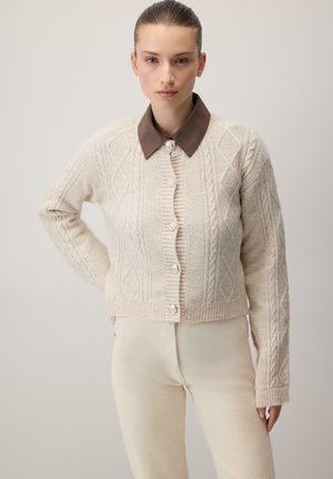 Kvinde iført en cremefarvet kabelstrikket cardigan med store knapper og mørkebrun krave, kombineret med cremefarvede bukser med høj talje, stående med hånden på hoften.