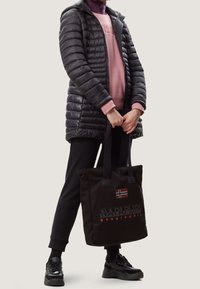Persona che indossa una giacca nera trapuntata, un felpa rosa, pantaloni neri e sneakers, mentre tiene una borsa tote nera con una bandiera e il logo del marchio.