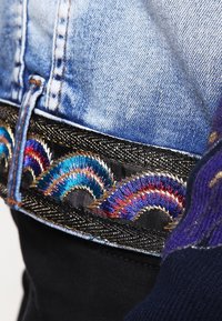 Denimtyg med livliga flerfärgade broderade vågor på ett svart midjeband, med metalliska trådar och texturförstärkningar.