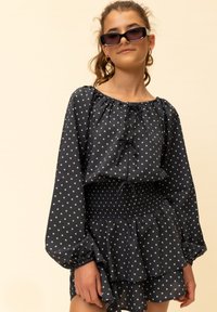 Marineblauwe jurk met wit sterpatroon, lange pofmouwen, een gathers halslijn en een gerimpelde taille met een ruches zoom. Gecombineerd met een zonnebril.