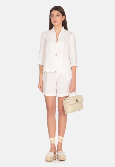 Blazer bianco in lino con una sottile trama, abbinato a pantaloni corti coordinati. Una borsa a tracolla beige imbottita e sandali con decorazioni completano il look.