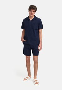Marinblå kortermet skjorte med lomme og matchende shorts, begge med strukturert stoff. Hvite sko fullfører antrekket.