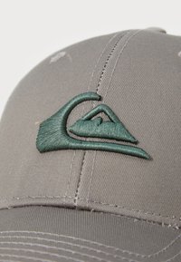 Grå baseballcaps med en grønn brodert logo, med en strukturert design, stikking detaljer og en jevn tekstur.