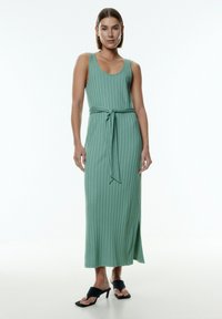 Mintgrünes, geripptes Maxikleid mit tailliertem Oberteil, Gürtel in der Taille und seitlichem Schlitz, kombiniert mit schwarzen, offen-toe High Heels.