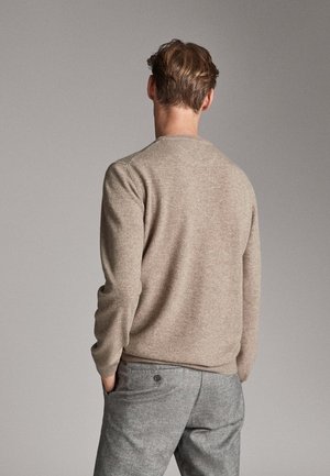 Homme aux cheveux bruns courts portant un pull beige et un pantalon gris, se tenant les mains dans les poches, de dos contre un fond neutre.