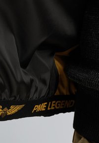 Čierna bomber bunda so žltým podšívaním, ktorá má textúrovaný pletený golier a manžety, a výrazný čierno-zlatý znak "PME Legend".