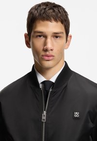 Black bomber jacket with a smooth finish, ribbed collar, and silver zipper. Features a small black logo patch on the chest.