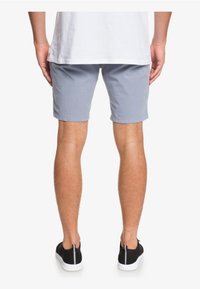 Osoba v světlých šedých šortkách, bílé tričko a černých šněrovacích teniskách stojí zády k bílé pozadí.