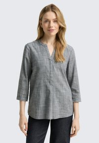 Mujer rubia vistiendo una blusa gris a cuadros con cuello en V y mangas 3/4, y pantalones oscuros, de pie contra un fondo gris claro.