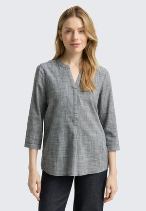 Donna bionda che indossa una blusa grigia a scacchi con scollo a V e maniche a 3/4, insieme a pantaloni scuri, in piedi contro uno sfondo grigio chiaro.