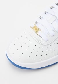 Valkoiset nahkakengät, joissa on rei'itetyt alueet hengittävyyden parantamiseksi. Mukana valkoiset nauhat, kultainen logo ja sininen kumipohja.