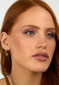 Nærbillede af en person med rødt hår, iført en gylden hoop ørestik med en klar accent, blød lyserød læbestift og subtil makeup i neutrale toner.