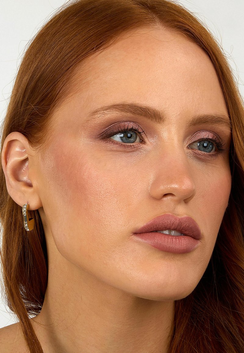 Nærbillede af en person med rødt hår, iført en gylden hoop ørestik med en klar accent, blød lyserød læbestift og subtil makeup i neutrale toner.