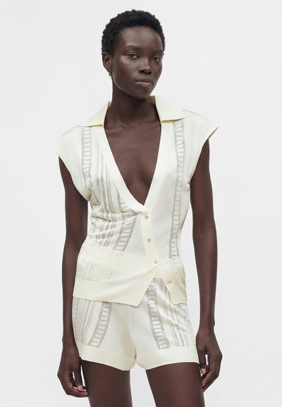 Γυναίκα με σκουρόχρωμο δέρμα που φοράει κρεμ πλεκτό κοντομάνικο τοπ με κουμπιά και αντίστοιχα σορτς, στέκεται μπροστά σε απλό ανοιχτό φόντο.