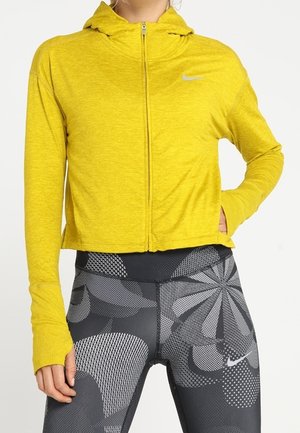 Kobieta ubrana w jaskrawożółtą kurtkę z kapturem na suwak z otworami na kciuki oraz czarne legginsy w abstrakcyjne szare i białe wzory.