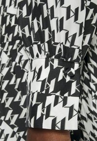 Černobílá látka s vzorem, obsahující ostré cik-cak tvary. Zahrnuje zapínání na knoflík a štítek s logem poblíž manžety.