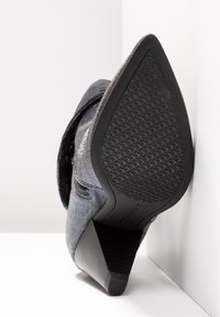 Botte à talon haut à bout pointu avec semelle en caoutchouc texturée et fermeture éclair latérale, inclinée vers le haut sur un fond d'angle blanc.