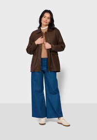 Kobieta w brązowej kurtce na beżowym swetrze, w niebieskich szerokich dżinsach i białych trampkach, stoi z rękami blisko zamka kurtki.