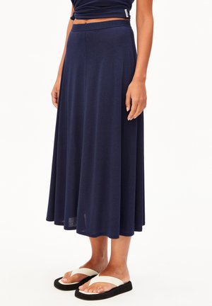 Frau trägt einen marineblauen Midirock und schwarze Flip-Flops mit weißen Riemen, steht vor einem weißen Hintergrund.