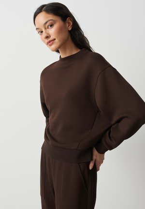 Donna con lunghi capelli scuri indossa una felpa marrone scuro e pantaloni coordinati, in piedi con una mano sul fianco davanti a uno sfondo semplice.