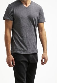 Uomo che indossa una maglietta a maniche corte V in grigio semplice e pantaloni neri, in piedi su uno sfondo bianco con le braccia rilassate.