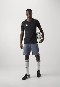 Schwarzes Adidas-T-Shirt mit kurzen Ärmeln und gelben Akzenten, graue Shorts, schwarze Fußballschuhe und weiße Socken mit schwarzen Streifen, hält einen Fußball.