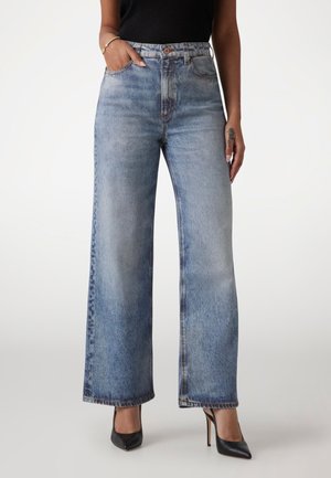 Frau trägt ausgewaschene, weit geschnittene blaue Jeans, schwarzes ärmelloses Oberteil, schwarze spitze High Heels, mit einer Hand in der Tasche und Tattoo an der anderen Hand.