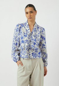 Vit blus med blå blommönster, knytdesign, krage och puffärmar; kombinerad med ljusgrå veckade byxor.