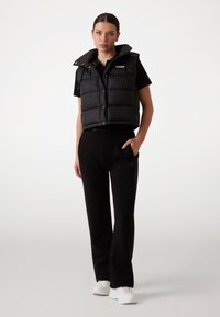 Gilet invernale nero con chiusura a bottone e colletto alto, indossato sopra una maglietta nera, abbinato a pantaloni neri e sneaker bianche.