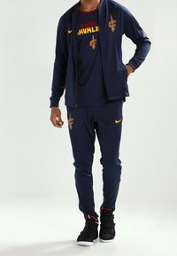 Mørkeblå atletisk træningsdragt med lynlåstrøje og bukser, med gule detaljer og Cleveland Cavaliers-logo. Sorte sporty sko med rødt detaljer.