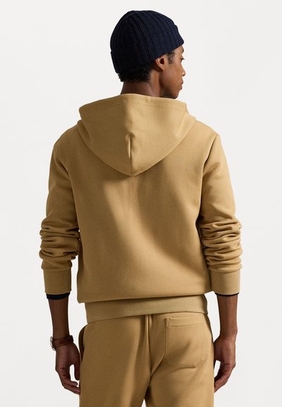 Sweatshirt com capuz em tom bege, com punhos e barra canelados, tecido texturizado e corte relaxado. Combinado com calças de jogging correspondentes, visto de costas. Gorro azul-marinho.