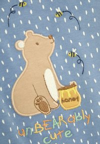 Design ricamato che presenta un orso con un vaso di miele su tessuto blu, accentuato da api gialle e pois bianchi. Il testo recita "incredibilmente carino."