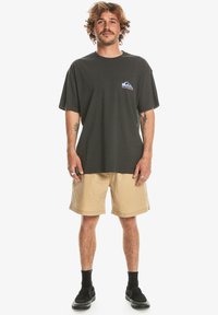 Camiseta de algodón negra con un pequeño logo azul y blanco en el pecho, combinada con pantalones cortos beige y zapatos negros sin cordones.