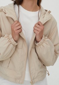 Chaqueta corta beige con puños elásticos y dobladillo ajustable. Presenta un cierre frontal de cremallera y dos bolsillos laterales. Usada sobre un sudadera blanca.