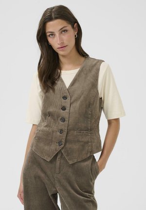 Brun fløjlsvest med V-hals, seks knapper og to forlommer, lagd over en cremet hvid t-shirt. Ensfarvet baggrund.