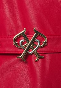 Červený kožený doplněk s propleteným logem v zlaté a stříbrné barvě. Hladký povrch s detailem prošívání podél okrajů.