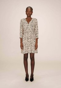 Robe beige avec un motif abstrait noir, décolleté en V, manches trois-quarts, taille froncée et jupe évasée. Chaussures noires à talons.