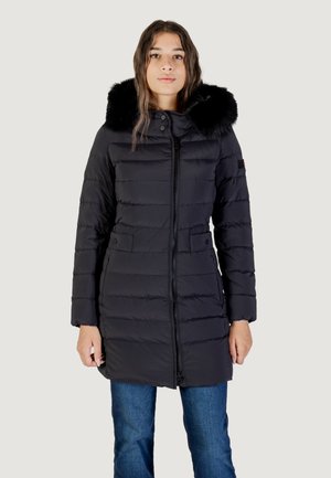 Donna che indossa un lungo cappotto nero trapuntato con cappuccio bordato di pelliccia nera, in piedi con le mani rilassate ai lati contro uno sfondo semplice.