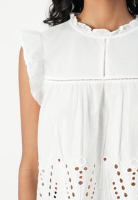 Blouse en coton blanc avec des accents de manches volantées, des détails de pintucks et des motifs découpés complexes le long de l'ourlet.