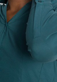 Maglietta teal con scollo a V e dettagli di pieghe sottili. Presenta un polsino con bottoni e una consistenza liscia. Inquadratura ravvicinata sull'area della spalla e sullo scollo.