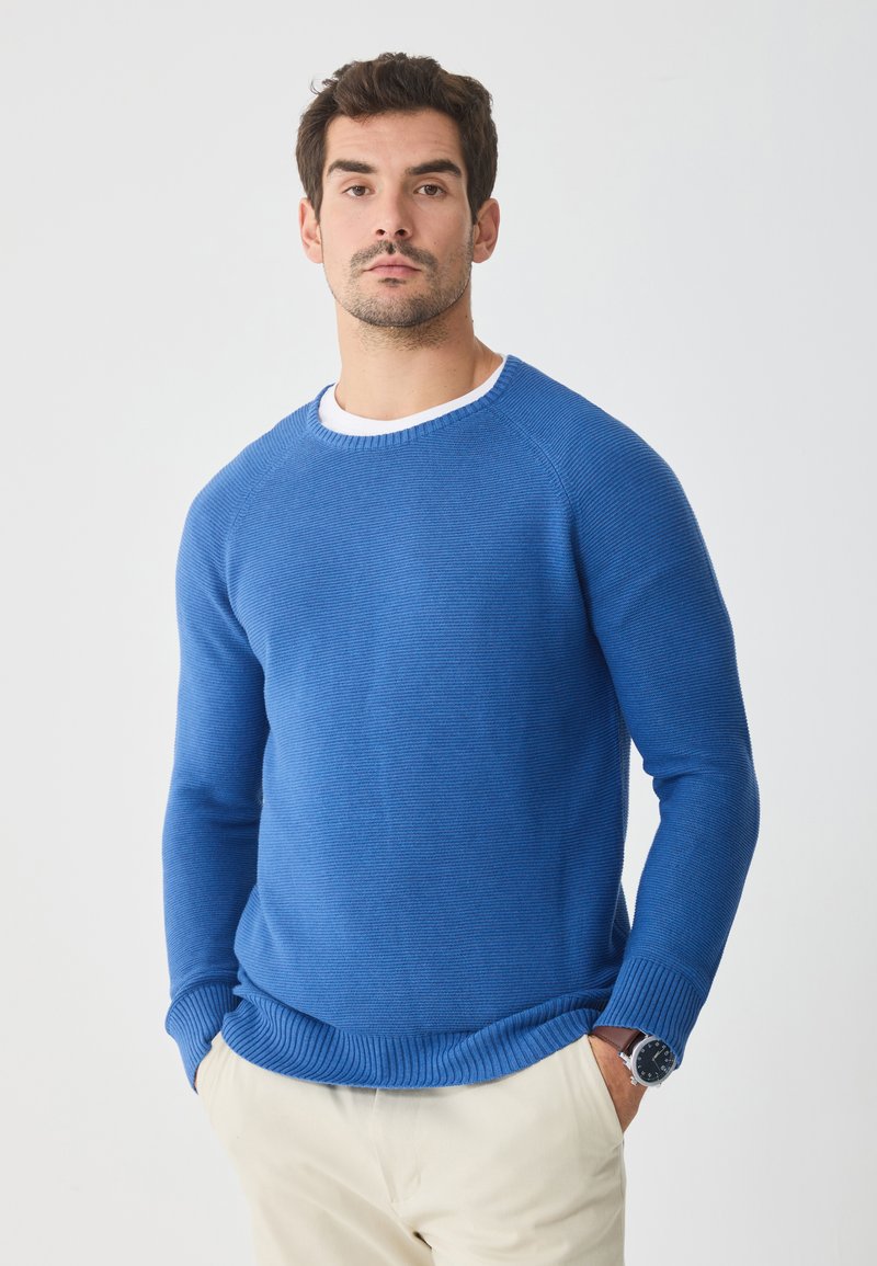 Blau gestreifter Strickpullover mit Rundhalsausschnitt, langen Ärmeln und strukturierter Oberfläche. Kombiniert mit hellen Hosen und einer Armbanduhr.